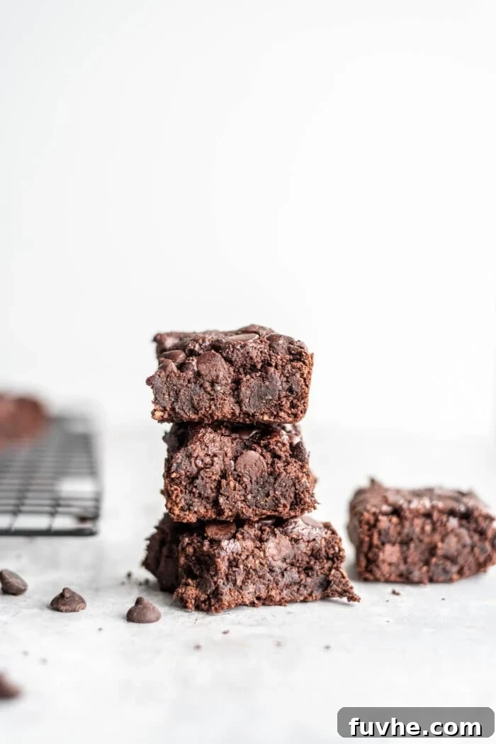 Stack of 3 vegan oat flour brownies on a grey surface with some chocolate chips scattered around, highlighting their fudgy texture.