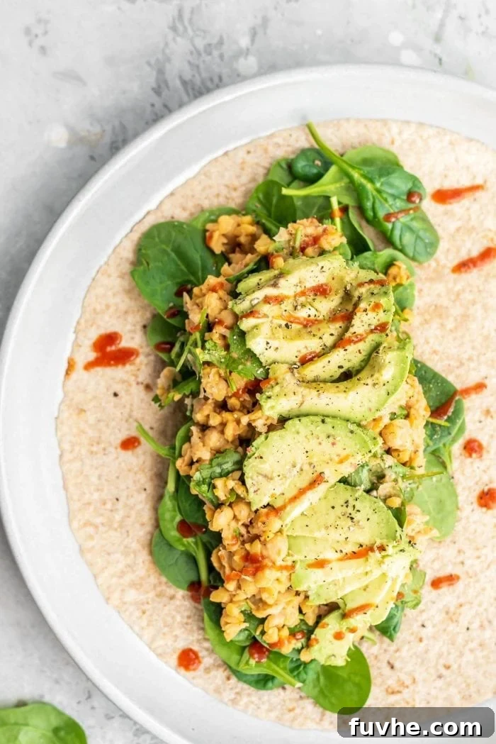 Kickin' Chickpea Avocado Wraps 5 A tortilla being assembled with a base of spinach, a scoop of spicy chickpea salad, avocado slices, and a final drizzle of hot sauce.