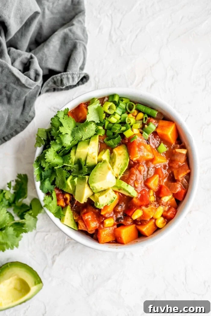 A vibrant bowl of homemade vegan sweet potato black bean chili, generously topped with creamy avocado slices, fresh cilantro, and finely chopped green onions, ready to be enjoyed.