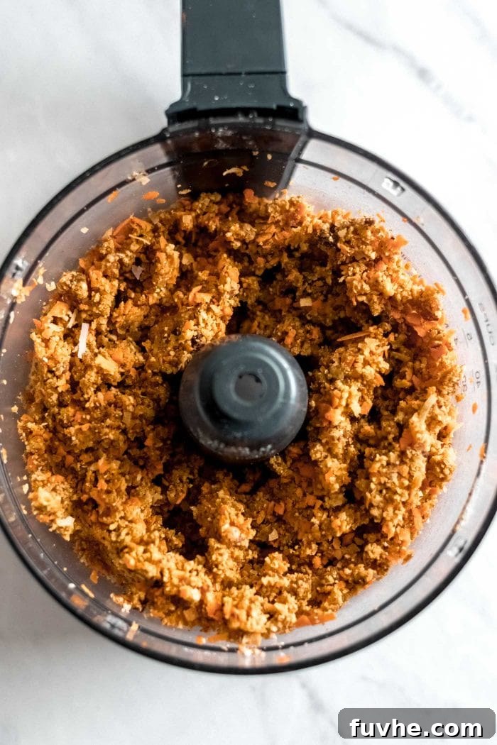 The blended dough for raw vegan carrot cake, a rich, moist mixture of nuts, dates, and carrots, collected in a food processor bowl.