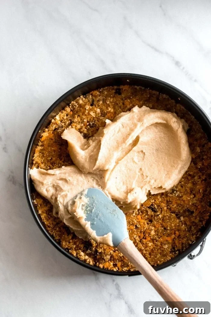 A springform cake pan holds the raw carrot cake base, as a spatula expertly spreads the thick cashew cream cheese frosting over its surface.