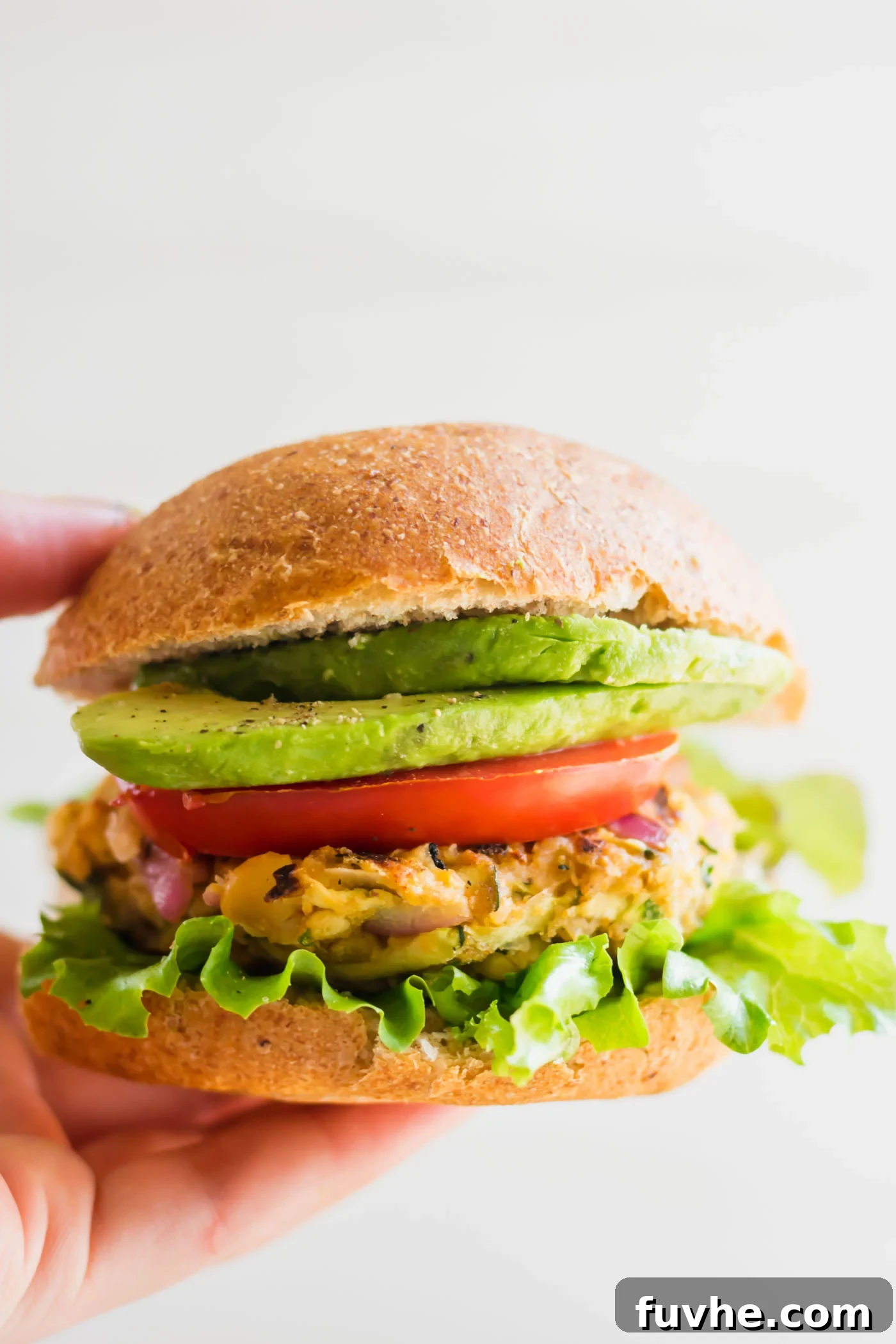Hand holding a chickpea veggie burger on a bun with lettuce, tomato, onion and avocado.