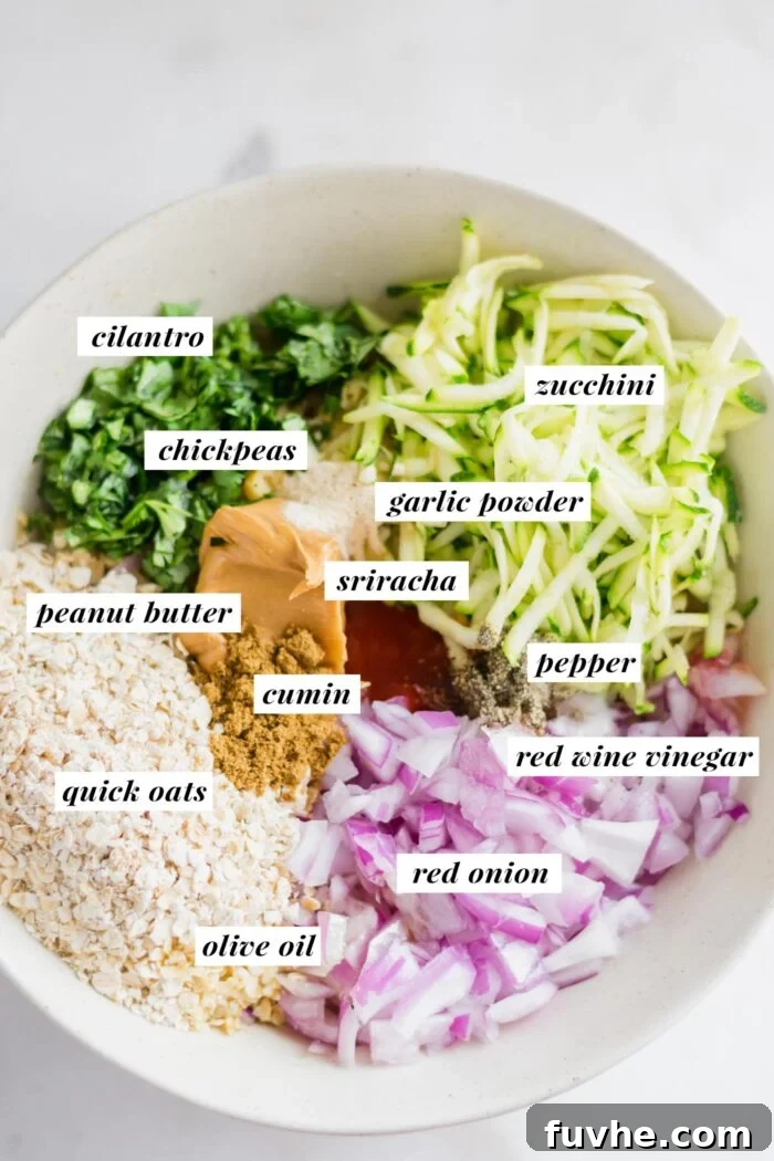 Large mixing bowl with grated zucchini, cilantro, red onion, oats, peanut butter and spices. Each ingredient is labelled with text.