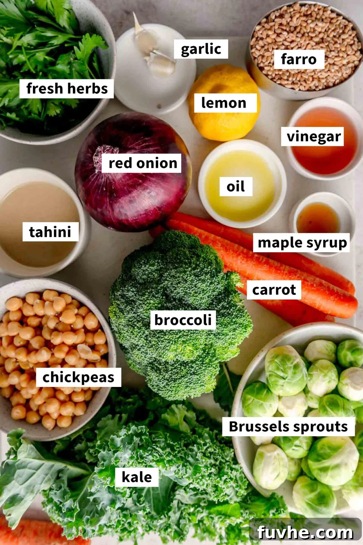 All the fresh ingredients for making a healthy vegan farro bowl, including tahini, fresh herbs, chickpeas, various vegetables, and farro, neatly arranged on a wooden cutting board with labels.