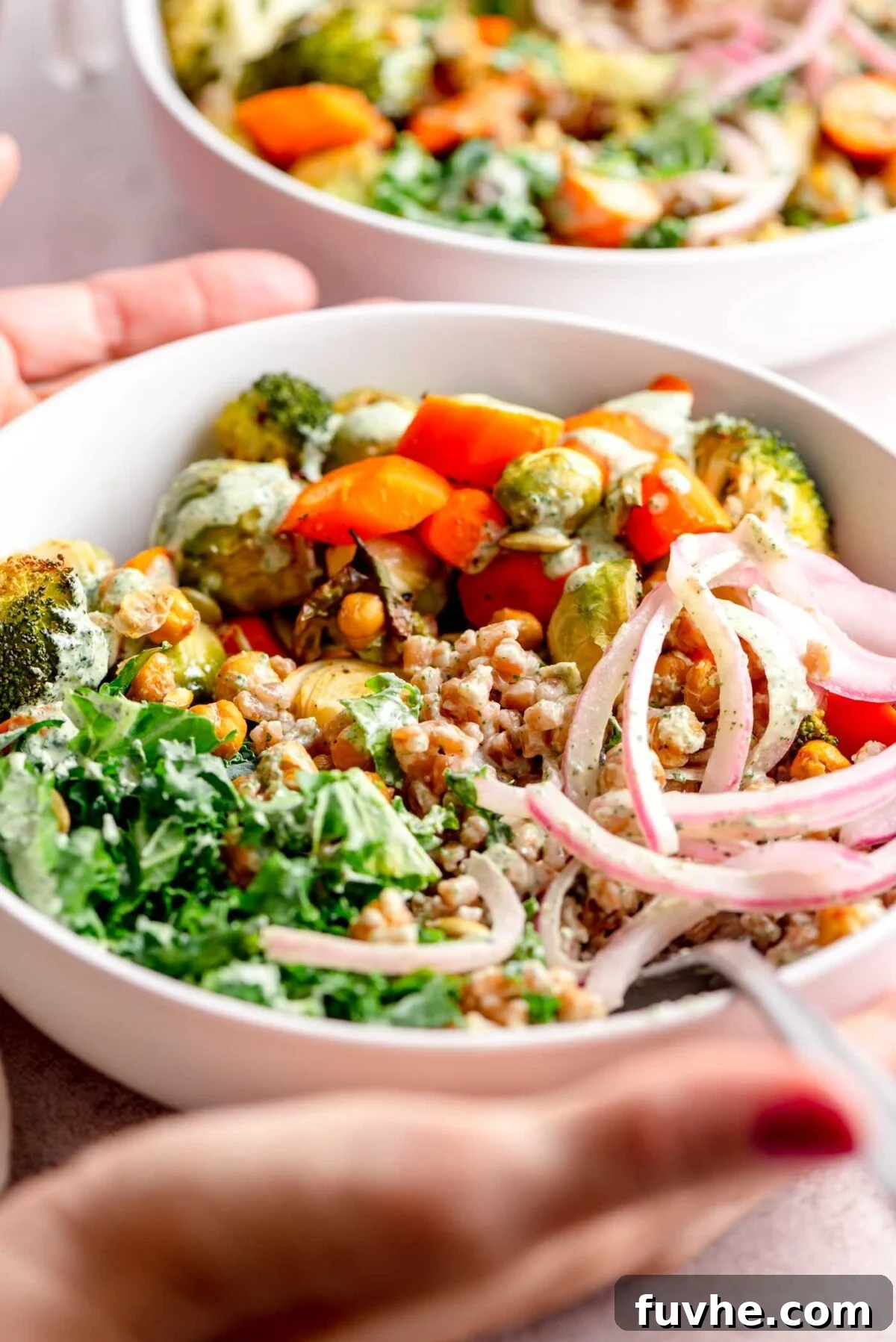 Hands holding a beautifully assembled vegan farro bowl with kale, roasted vegetables, crispy chickpeas, pickled onion, and a generous drizzle of creamy sauce.