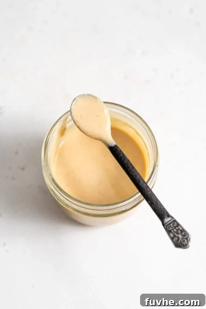 A spoon expertly scooping out a generous portion of creamy miso tahini dressing from a small glass jar, emphasizing its luxurious texture.