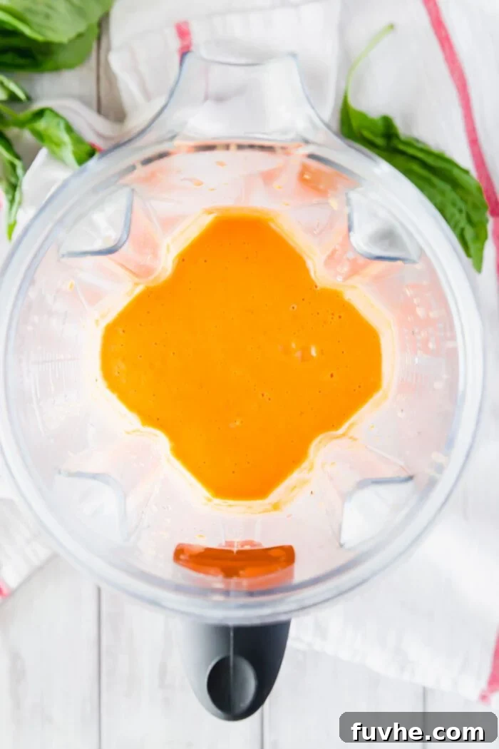 A high-speed blender container filled with roasted red peppers, cannellini beans, nutritional yeast, and almond milk, ready to be blended into a creamy sauce.