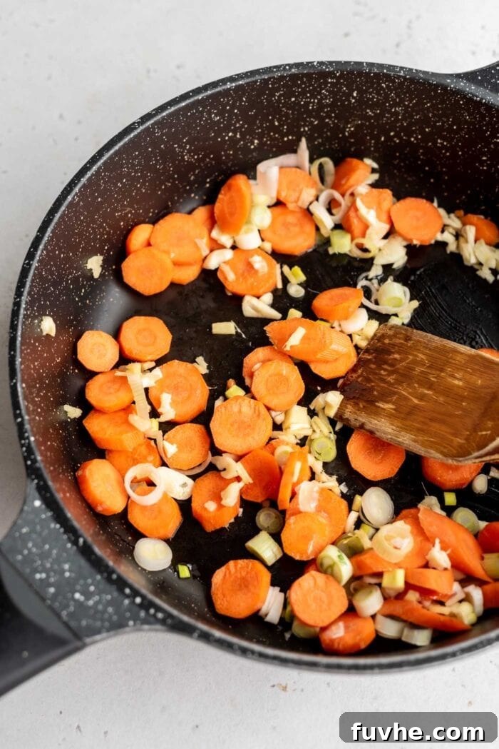 Carrots, ginger, garlic, and the white parts of scallions sautéing in a skillet with a wooden spoon, releasing their aromas.