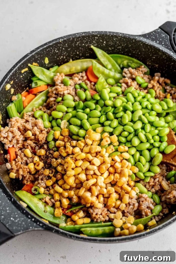 Cooked farro, corn, edamame, carrots, and snow peas are combined in a skillet, ready to be stir-fried.