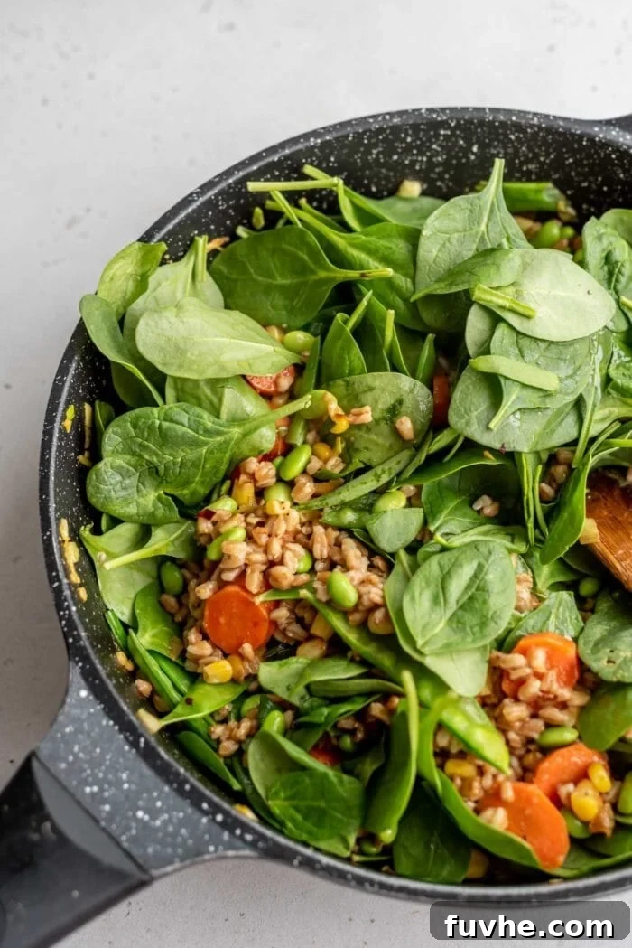 Fresh baby spinach being mixed into the hot farro and vegetable stir-fry in a skillet.