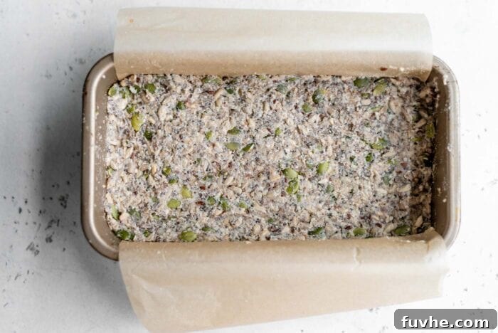 The raw seed bread dough, neatly pressed into a parchment paper-lined loaf pan, poised and ready to be placed in the oven for baking.