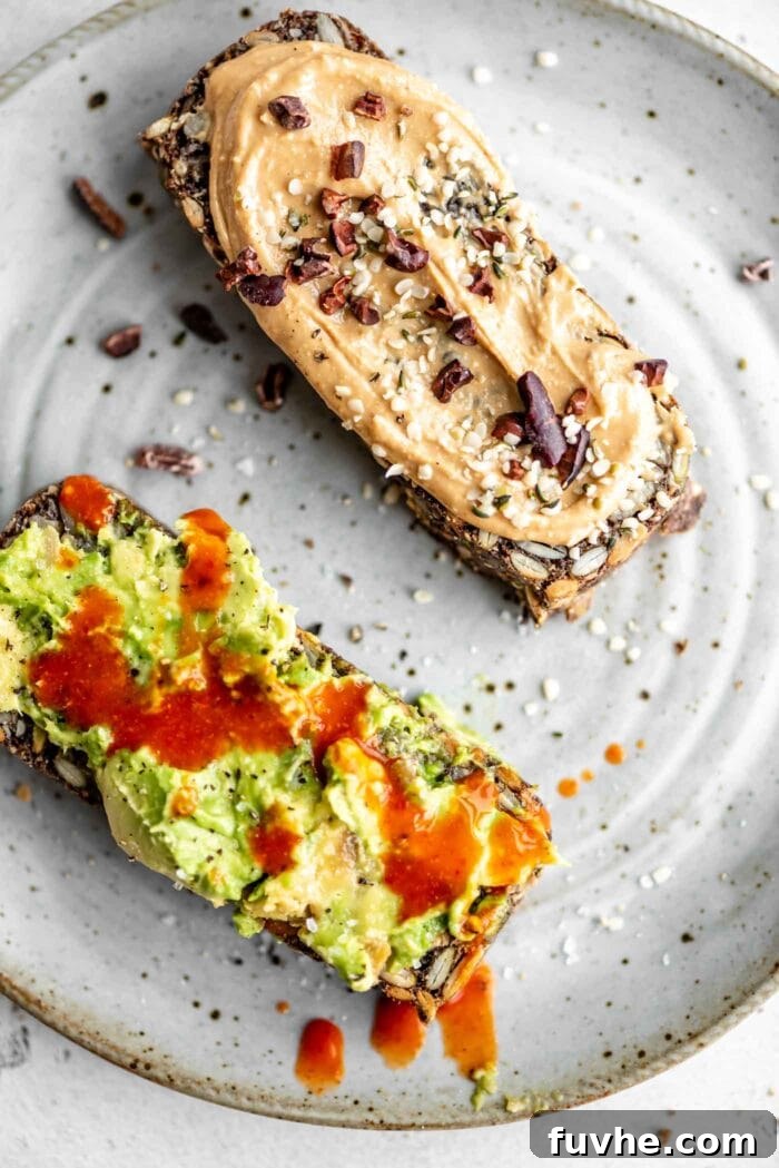 Two exquisite slices of homemade seed loaf presented on a small white plate; one slice is generously topped with creamy avocado, while the other features a delightful spread of peanut butter sprinkled with crunchy cacao nibs, showcasing versatile serving suggestions.