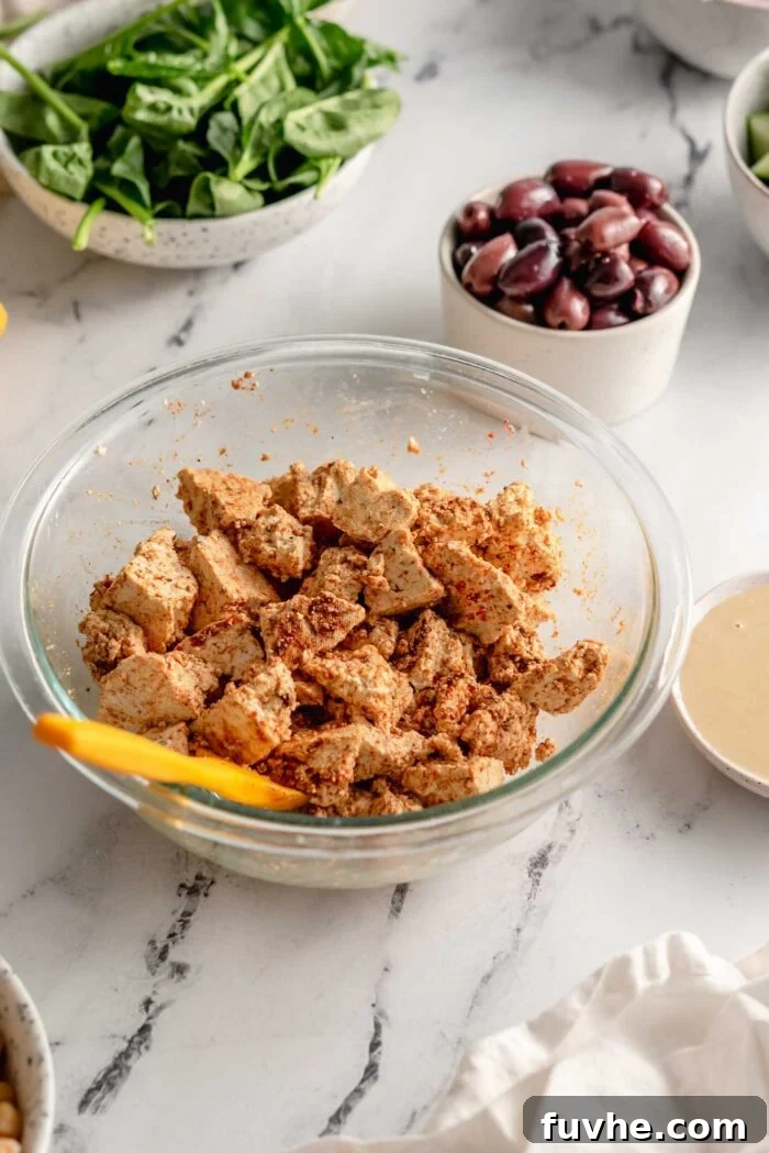 Irregularly torn pieces of firm tofu in a mixing bowl, thoroughly coated with a blend of aromatic shawarma spices, with a small spatula, olives, and spinach in the background.