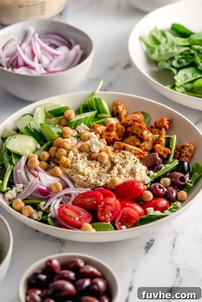 An exquisitely arranged healthy bowl brimming with fresh spinach, crispy shawarma tofu, creamy hummus, vibrant red onion slices, juicy tomatoes, briny olives, and whole chickpeas. Smaller ingredient bowls are alongside for visual context.