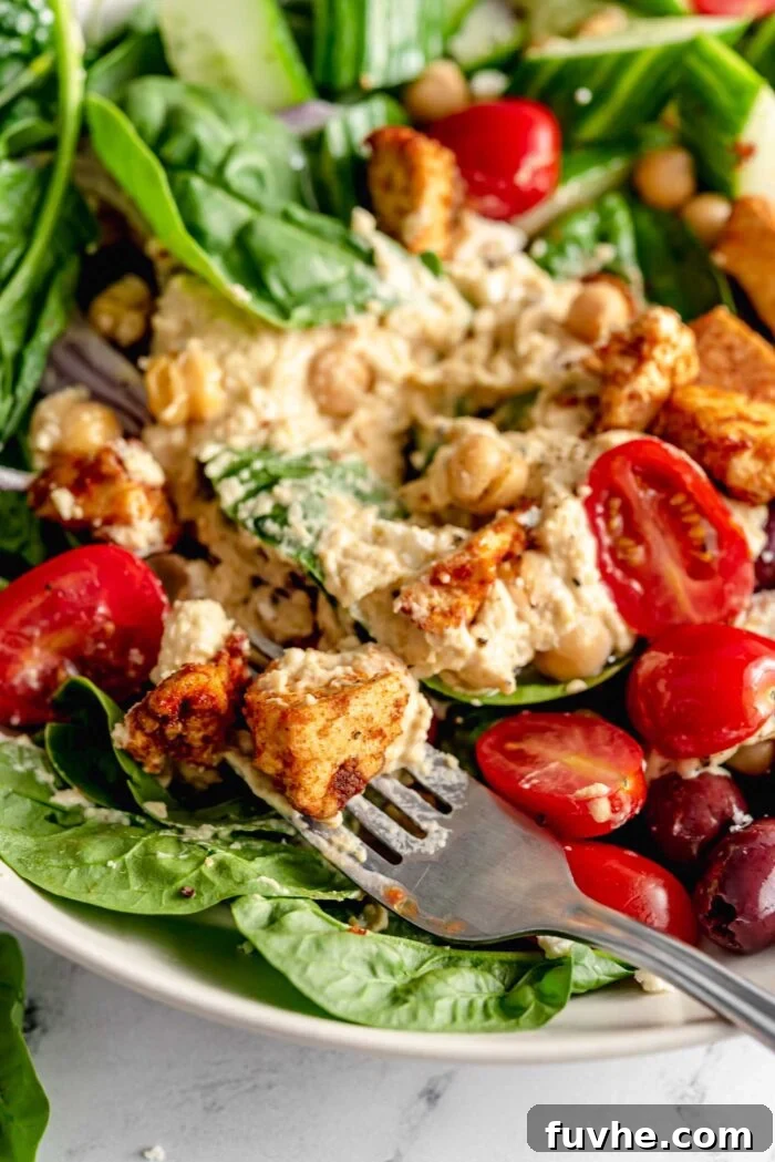 A fork piercing into a delicious Mediterranean hummus bowl filled with fresh spinach, juicy tomatoes, briny olives, crispy shawarma tofu, creamy hummus, and chickpeas, highlighting the inviting textures.