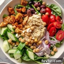 Overhead view of a colorful and healthy bowl featuring fresh spinach, creamy hummus, halved tomatoes, sliced red onion, crisp cucumber, and olives, a perfect meal.