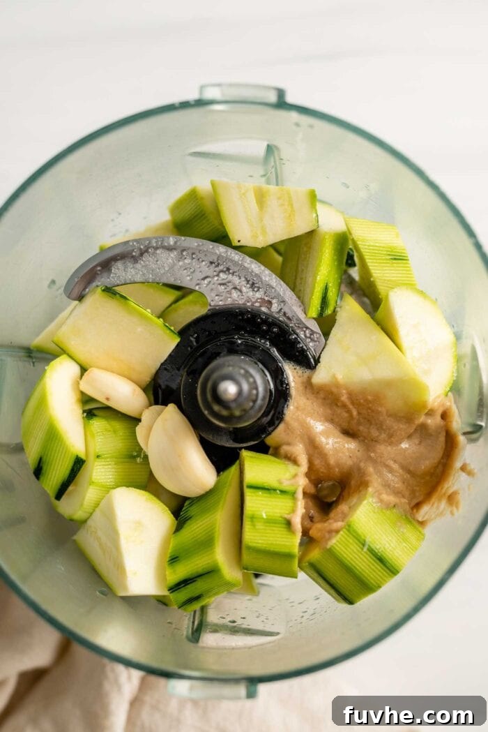 Roughly chopped zucchini, whole garlic cloves, and creamy tahini are combined in a food processor, poised to be blended into a smooth dip.