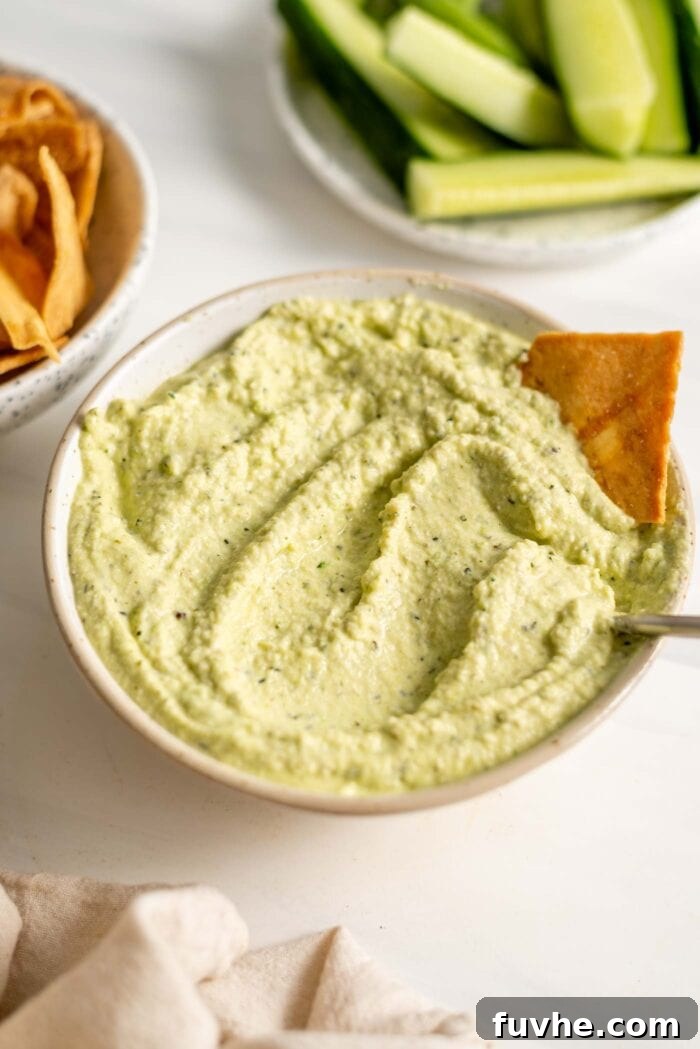 A beautifully presented bowl of vibrant green zucchini dip, with a spoon nestled inside and a golden pita chip invitingly dipped into its creamy texture.