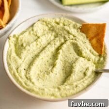 A bowl of vibrant green zucchini dip, garnished with fresh herbs and served with crisp cucumber slices and golden pita chips.