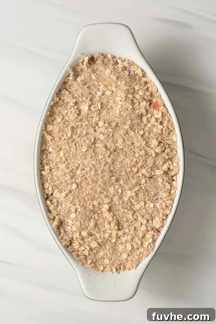 An uncooked oat crumble topping spread over the fruit filling in an oval baking dish, ready for the oven.