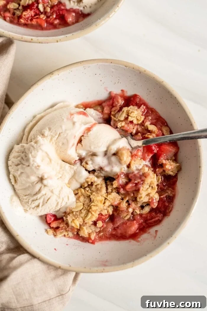 A beautifully presented bowl of vegan strawberry rhubarb crisp, topped with a melting scoop of vanilla ice cream, and a spoon ready for digging in.