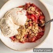 A bowl of vegan strawberry rhubarb crisp with vanilla ice cream and a spoon in it.
