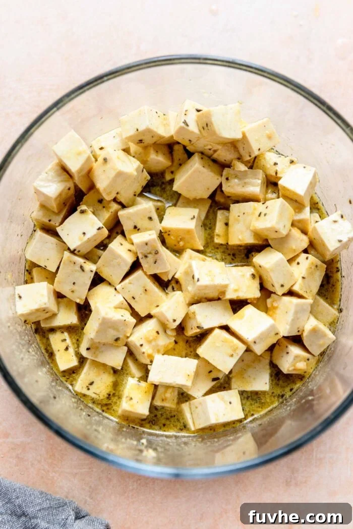 Cubes of firm tofu marinating in a flavorful blend of herbs, lemon juice, and olive oil, resting in a clear glass bowl, absorbing all the aromatic goodness.