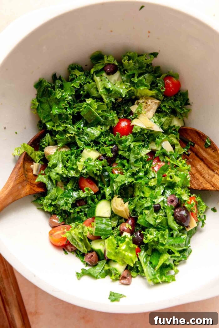 A large mixing bowl filled with massaged kale, ripe tomatoes, marinated artichoke hearts, briny olives, and crisp cucumber, with two wooden serving spoons ready to toss the salad.