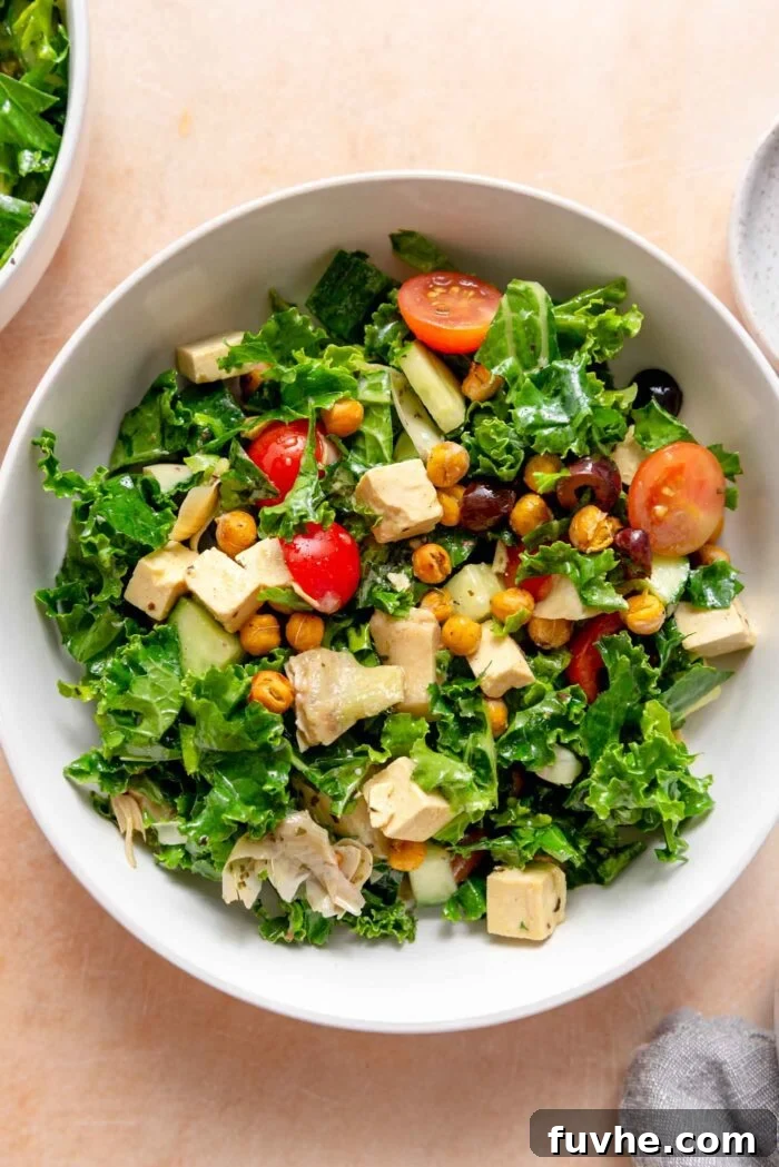 A vibrant bowl of Mediterranean kale salad, garnished with chopped tomatoes, artichoke hearts, fresh cucumber, crunchy roasted chickpeas, savory tofu feta cubes, and chopped Kalamata olives.