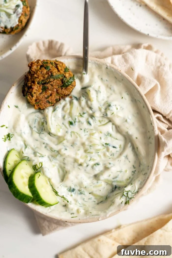 A bowl of creamy, white vegan tzatziki with chopped dill, garnished with a falafel and a few slices of fresh cucumber, invitingly ready to be served.