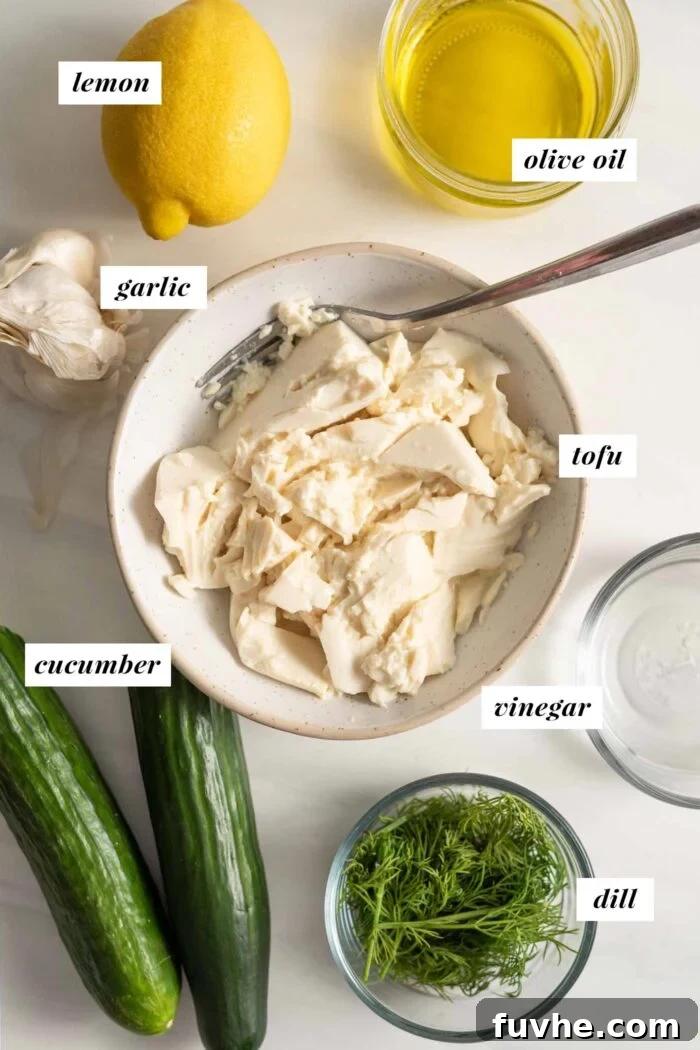 A vibrant flat lay of fresh ingredients including a block of soft tofu, whole lemons, cloves of garlic, a crisp English cucumber, fresh dill sprigs, and a bottle of olive oil, ready for making vegan tzatziki.