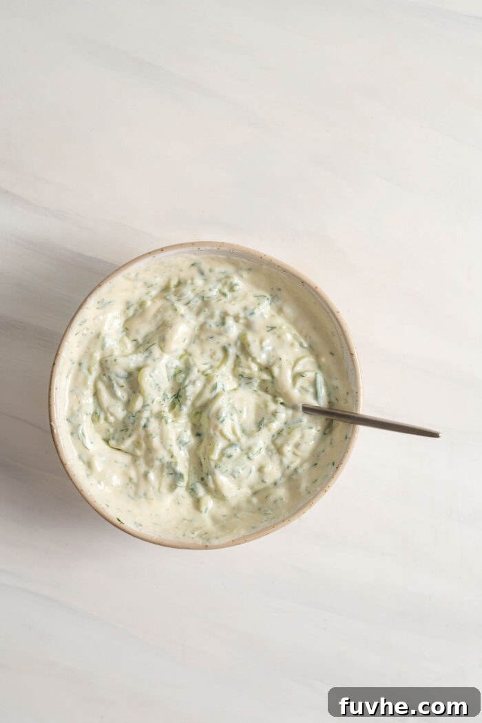 A bowl of freshly made creamy vegan tzatziki dip with a spoon, showcasing the perfect texture and the flecks of green dill throughout.