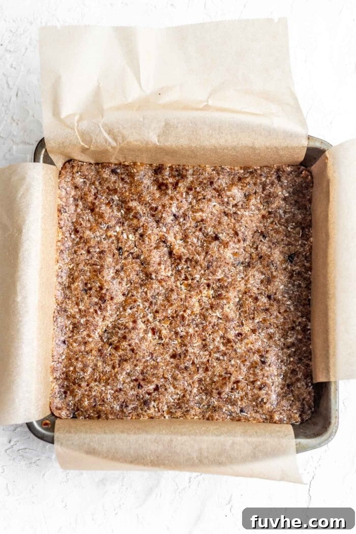 Decadent Plant-Based Chocolate Coconut Squares 5 Raw coconut date bars pressed into a baking pan lined with parchment paper, showcasing the even base before adding chocolate.