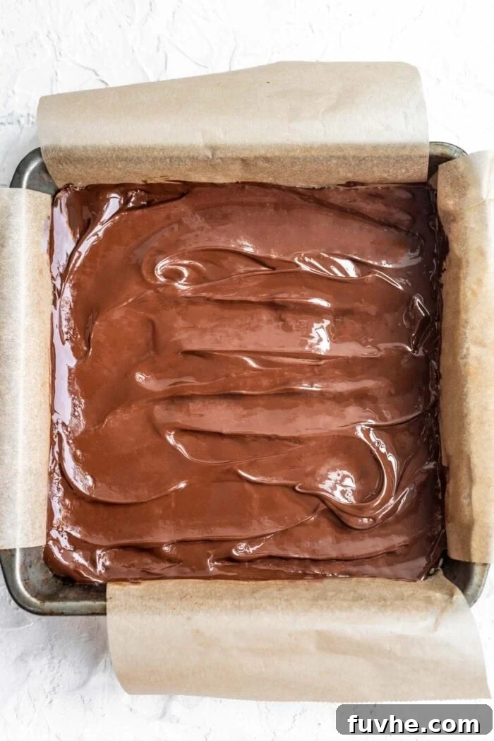 Decadent Plant-Based Chocolate Coconut Squares 7 Melted dairy-free chocolate being spread evenly over the firm coconut base in a baking pan, creating a smooth top layer.