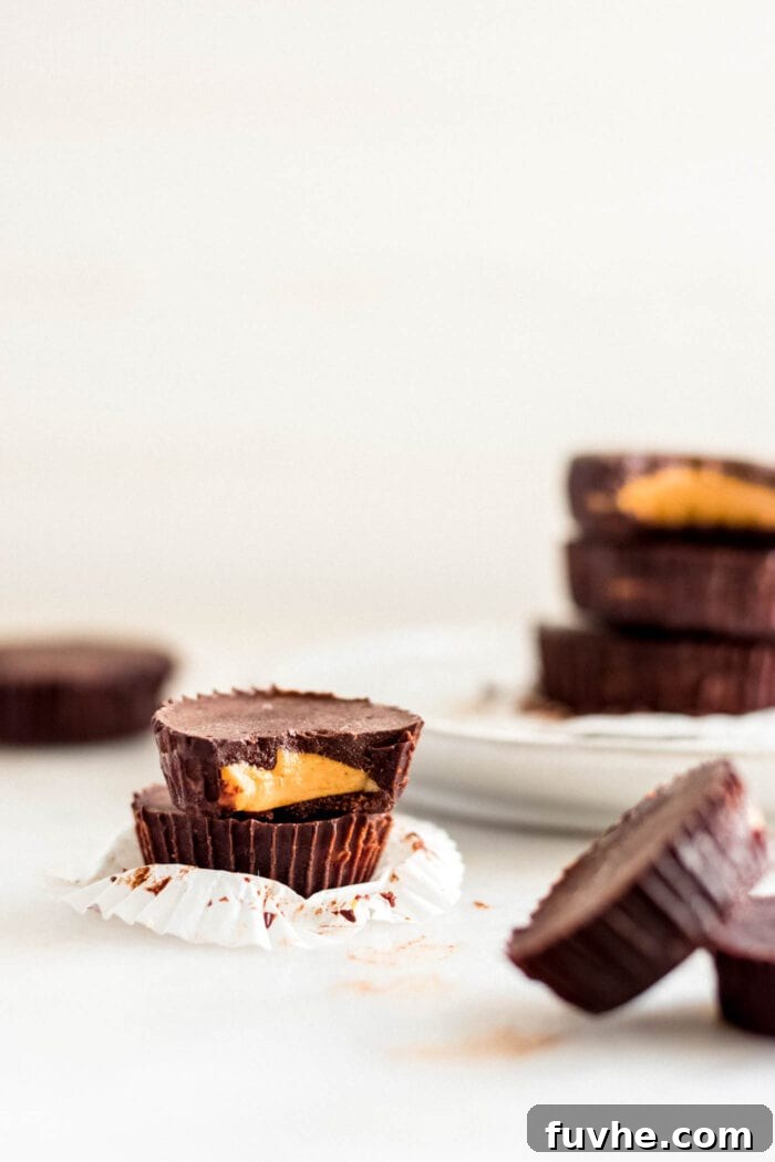 Two perfectly stacked vegan chocolate peanut butter cups, one with a bite taken out revealing the creamy filling, with more cups blurred in the background.