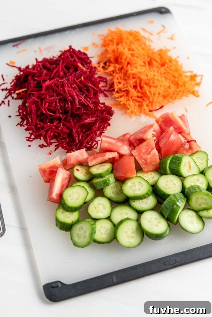 A cutting board laden with finely chopped cucumber, diced tomato, and vibrant grated beet and carrot.