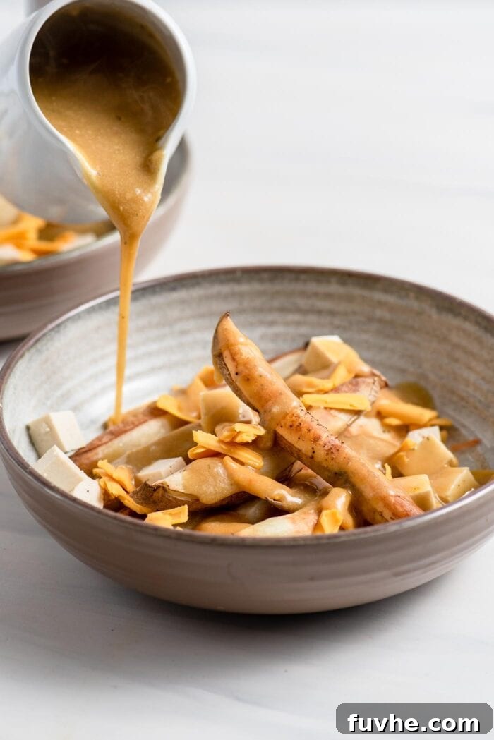 Thick, creamy miso gravy being poured over a base of golden baked fries, cubed tofu, and grated vegan cheese in a serving bowl.