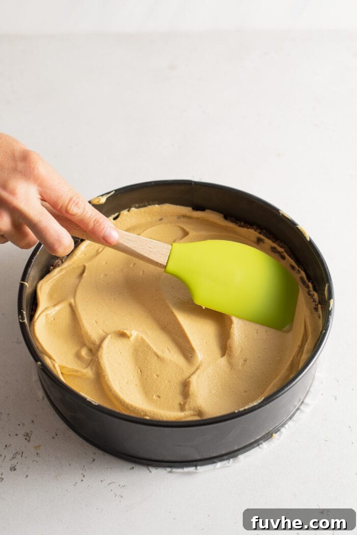 Easy Vegan No-Bake Pumpkin Cheesecake 7 A spatula gently spreading the smooth pumpkin cheesecake filling into the prepared date and walnut crust within a springform pan.
