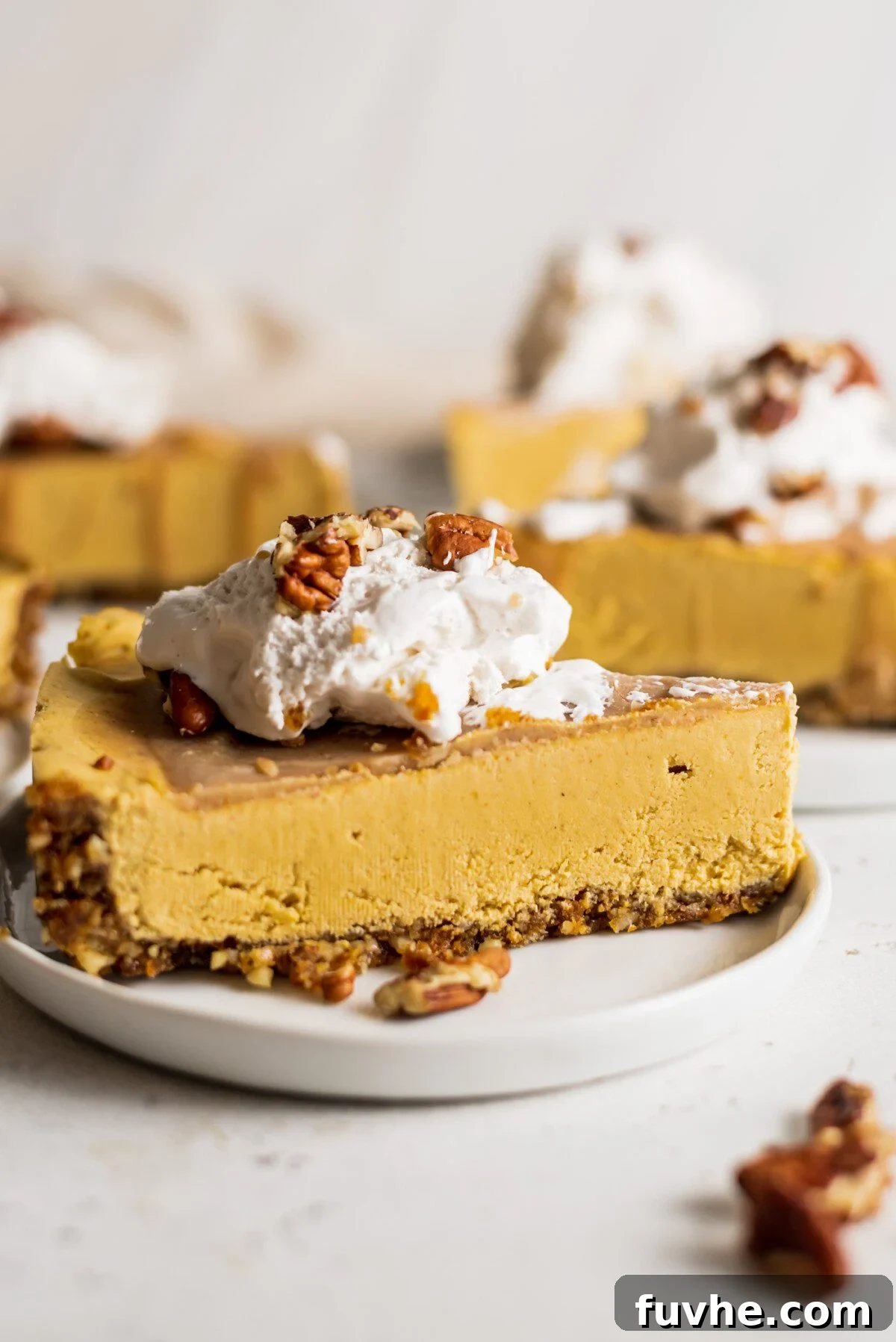 Easy Vegan No-Bake Pumpkin Cheesecake 8 A beautifully presented slice of vegan no-bake pumpkin cheesecake adorned with whipped cream and candied pecans on a plate, with more slices visible in the background.