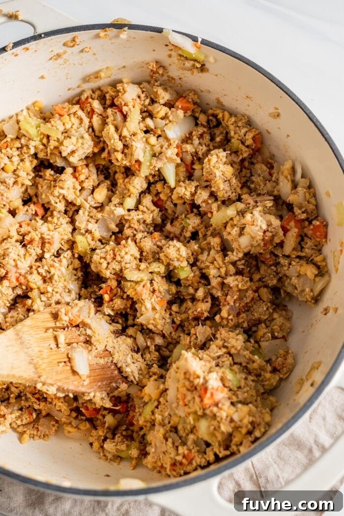 The uncooked chickpea loaf mixture, a rich blend of vegetables, oats, and chickpeas, is thoroughly combined in a large pot, with a wooden spoon resting in it.