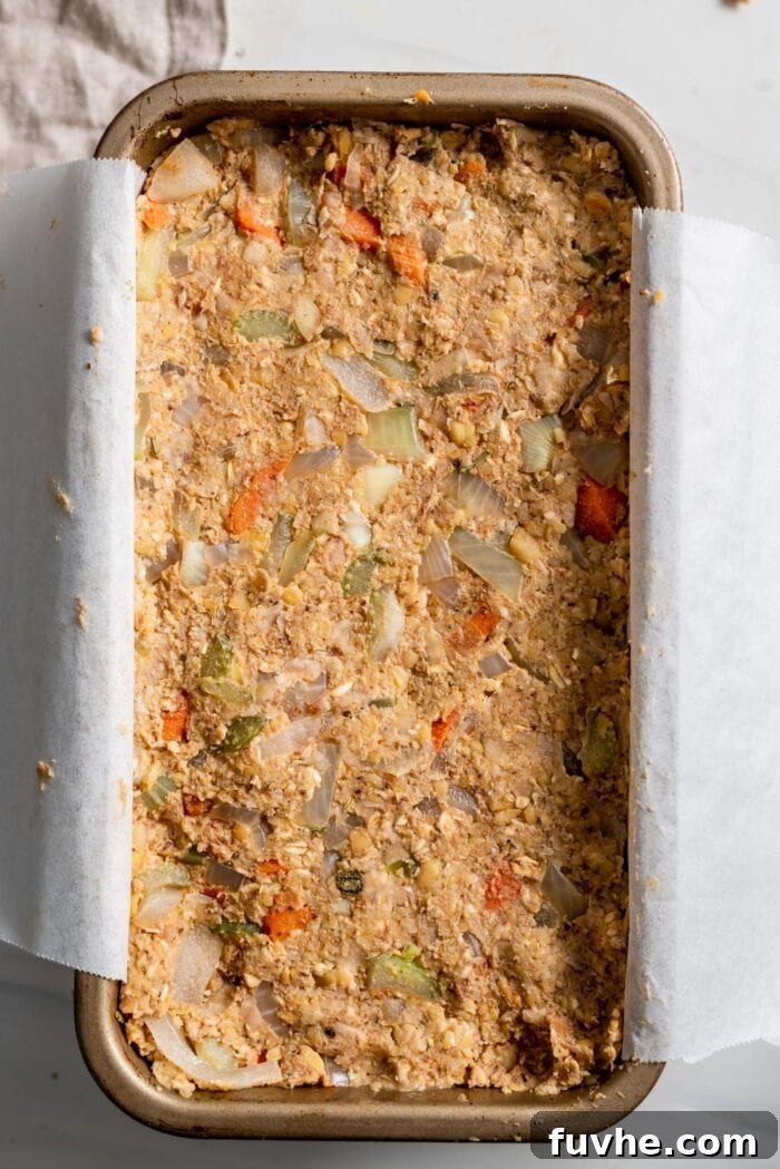 The unbaked chickpea loaf mixture is firmly pressed into a loaf pan, carefully lined with parchment paper, awaiting its flavorful glaze and trip to the oven.