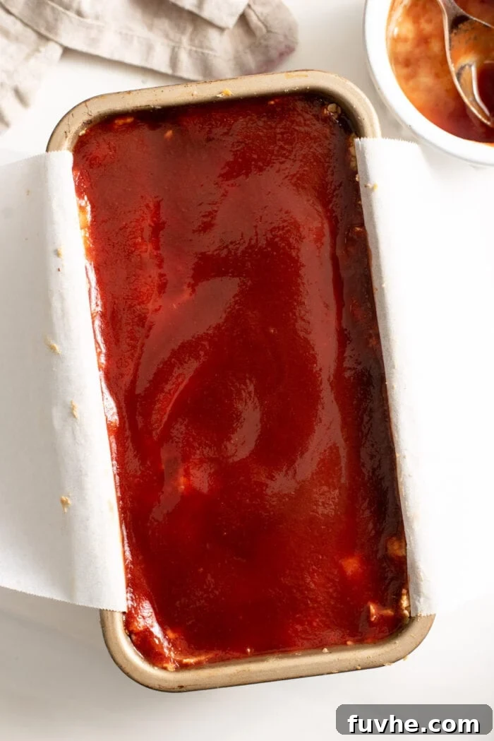 The raw vegan loaf, nestled in its parchment-lined pan, is generously topped with a rich, glossy tomato glaze, promising a delicious and flavorful crust.