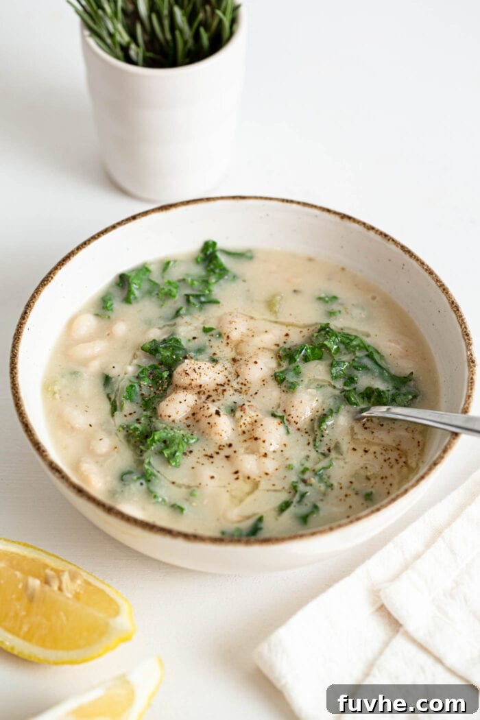 Nourishing Tuscan Kale and White Bean Soup 8 Bowl of white bean and kale soup with a spoon in it.. Sliced lemon rests in foreground.