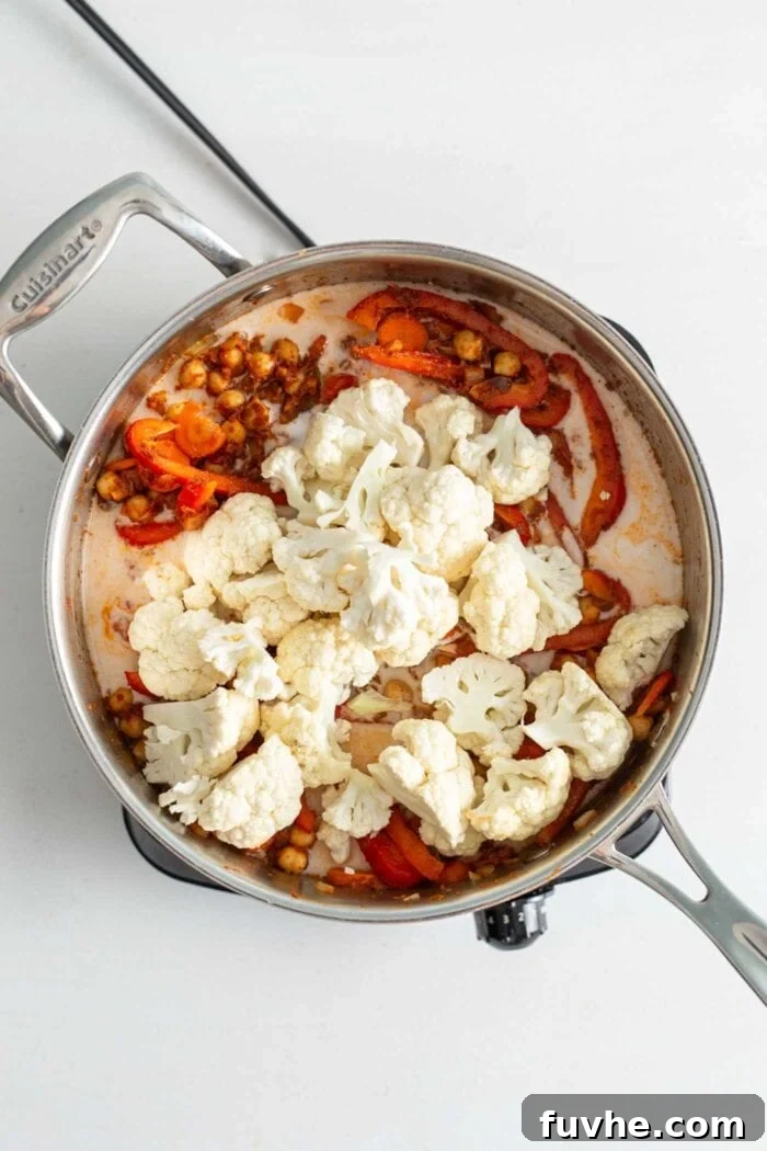 Chickpeas, bell peppers, cauliflower and carrots cooking in coconut milk in a skillet on a small cook top.
