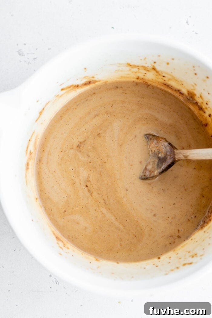 All wet ingredients for healthy blueberry muffins, including mashed banana, almond butter, coconut sugar, vanilla, plant milk, and lemon juice, mixed smoothly in a bowl.