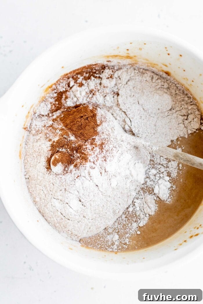 Dry ingredients, including whole wheat flour, baking powder, cinnamon, and salt, are gently mixed into the wet ingredients in a large bowl, forming the muffin batter.