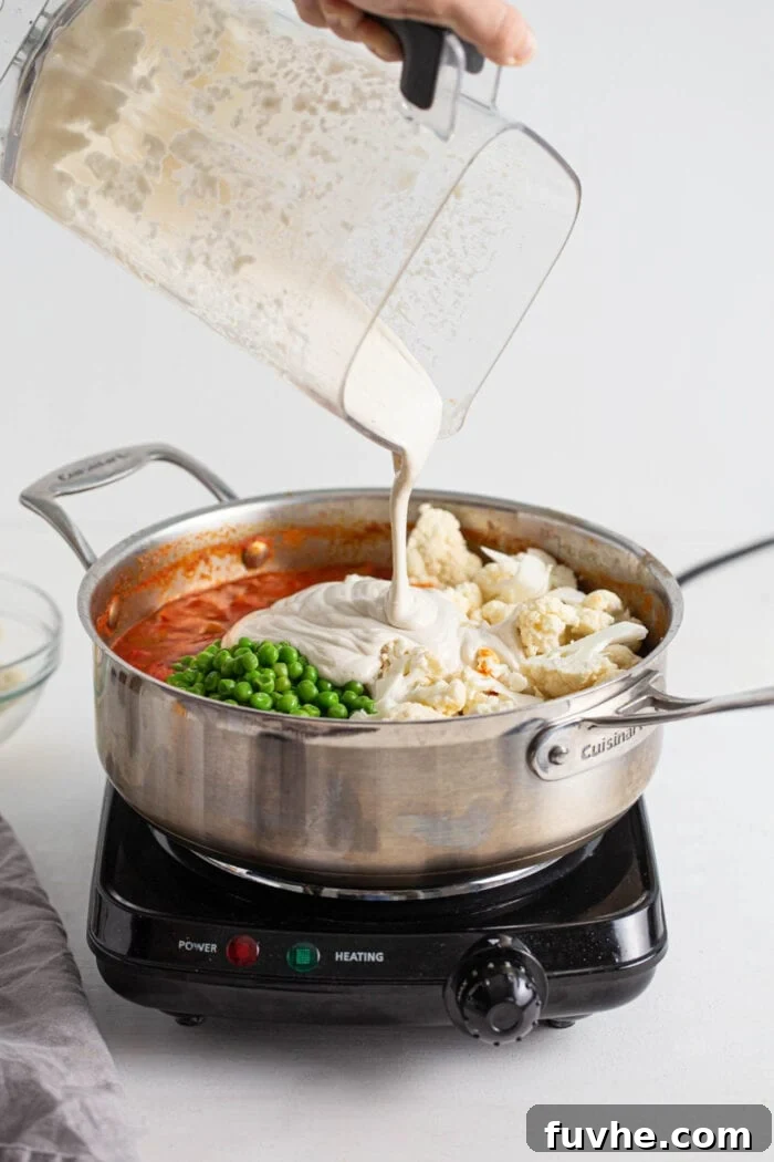Creamy Vegan Vegetable Korma 9 A blender pouring the silky cashew cream into the simmering vegetable korma, which includes cauliflower florets and green peas, enriching the sauce.