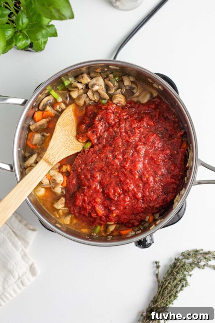Hearty Vegan Mushroom Bolognese 6 Crushed tomatoes in a skillet with carrots, celery and diced mushrooms. Wooden spoon rests in skillet.