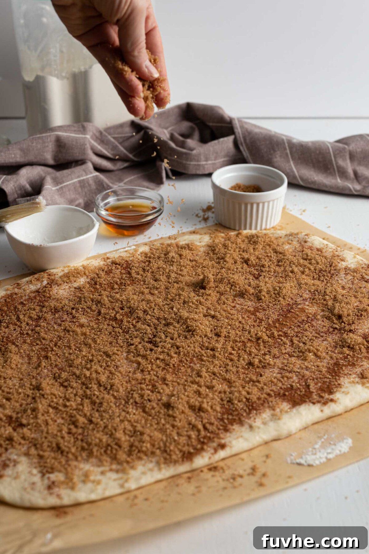 Hands sprinkling a generous amount of brown sugar and cinnamon over a rolled-out cinnamon roll dough rectangle.