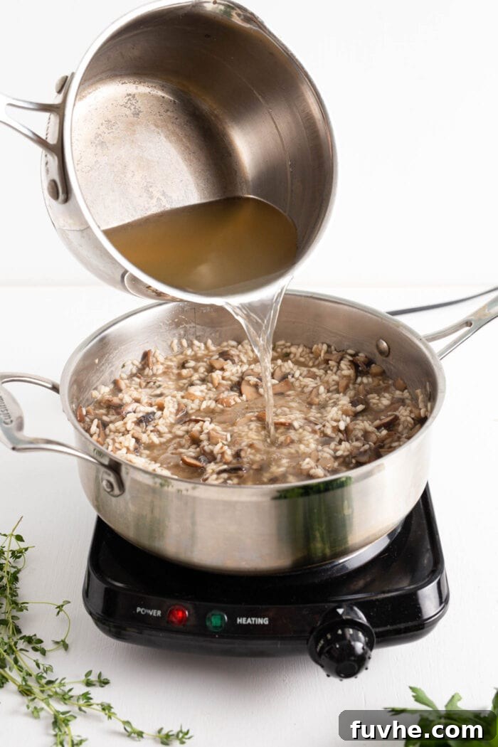 A continuous stream of hot vegetable stock being slowly poured from a measuring cup into a skillet containing rice and mushrooms, as the risotto begins to cook.